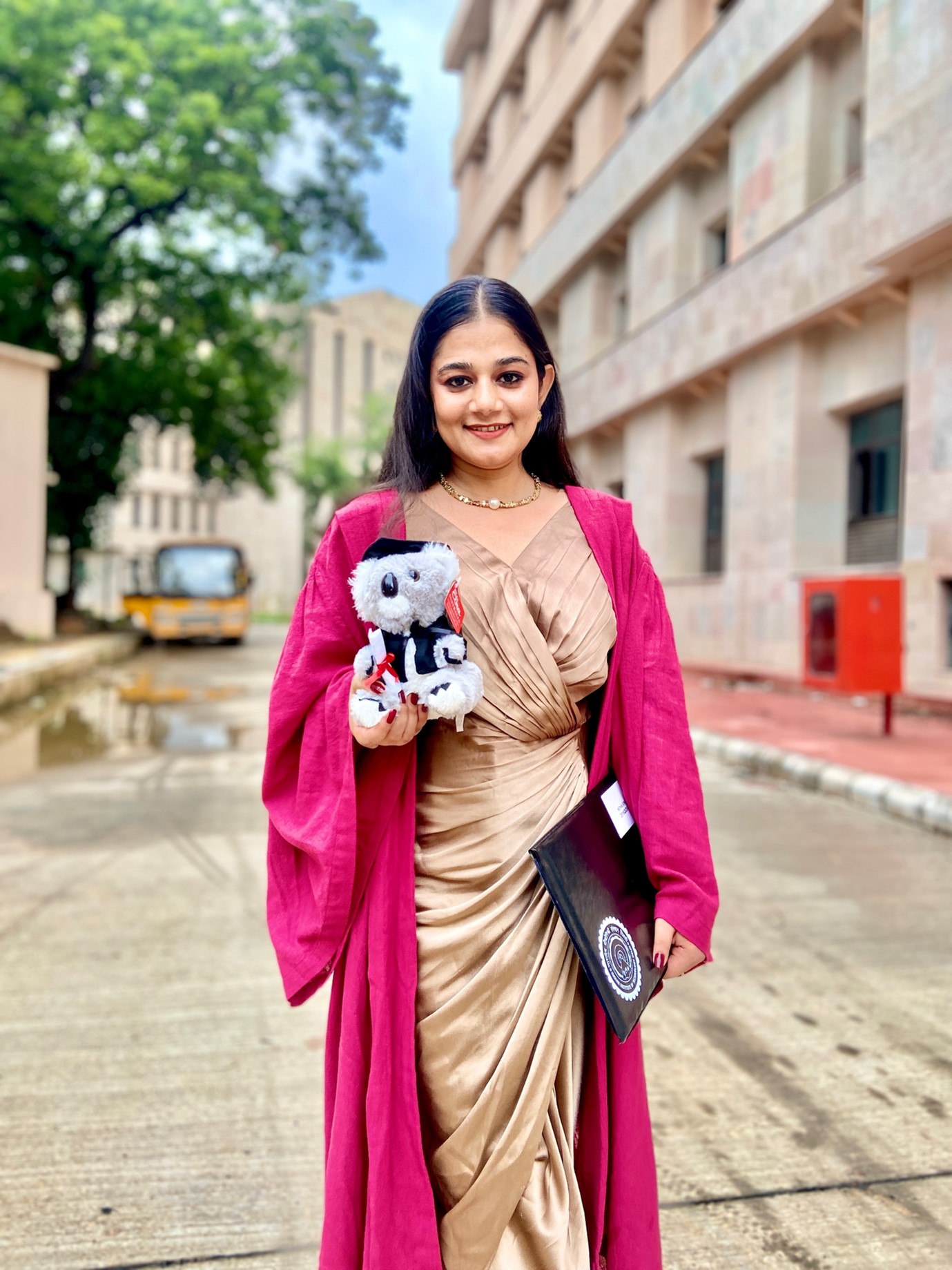 Dr Neha Singh standing in front of a building wearing a pink graduation robe.