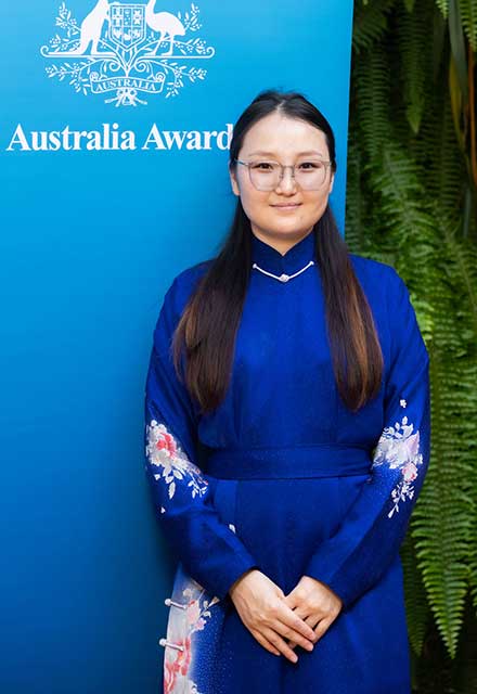 Dr  Ambaselmaa Bayarsaikhan standing in front of an Australia Awards banner wearing a blue dress.