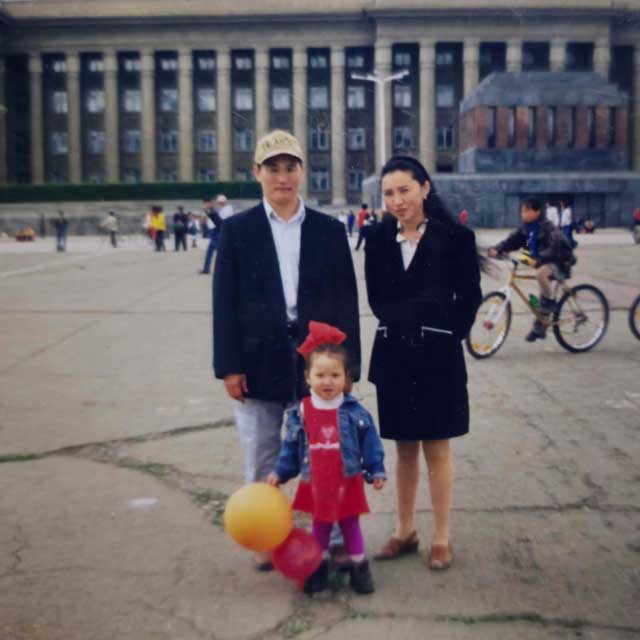 Dr  Ambaselmaa Bayarsaikhan as a child, standing with her parents in a public square in Mongolia. She wears a red dress, has a large red ribbon in her hair, and holds two balloons.