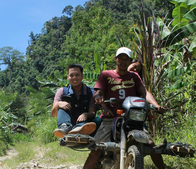 Dr Manalo on the side of a motorbike with colleague getting taken to remote school and farm