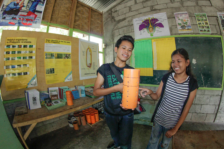 students (male and female) demonstrating importance of monitoring water levels in rice fields - they're inside a grass thatched roof