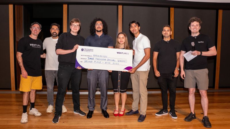 Taha holding UQ Ventures check for second place on stage with team mates - smiling and happy