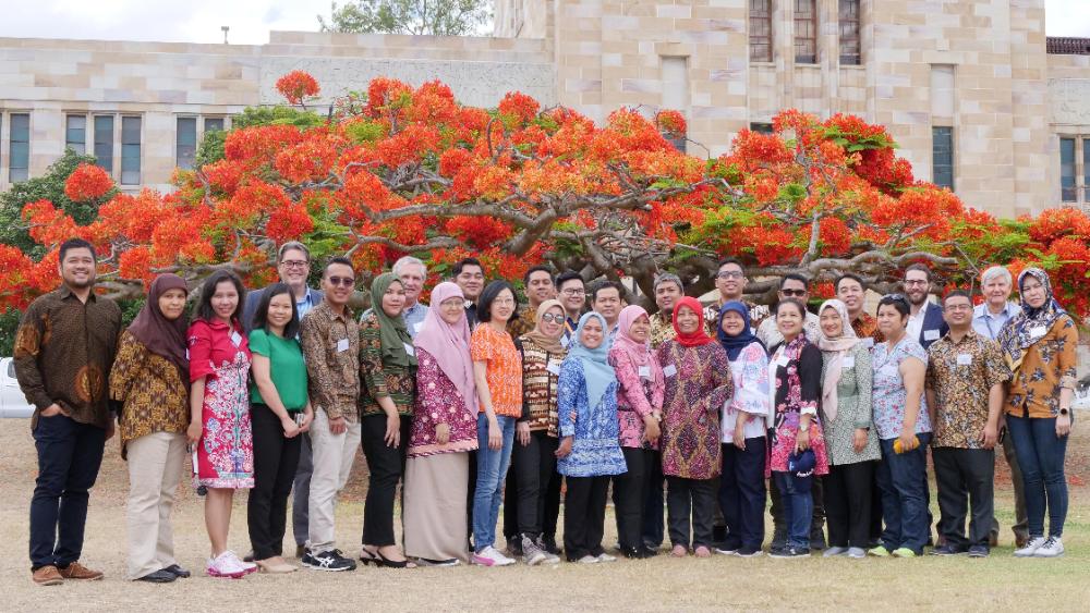 Australia Awards participants gathered as one group standing in front of a flame red tree on the grounds of the Great Court, St Lucia UQ 