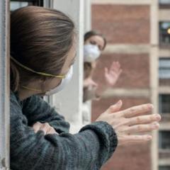 two women waving to one another from adjacent balconies - both wearing masks