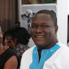 Richard Ellimah sitting in a conference audience - well lite - smiling to camera wearing a white shirt with a blue collar