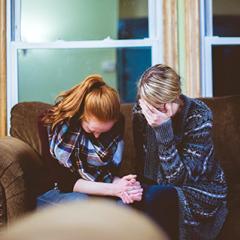 Two women head down on a couch with glass windows behind them - appearing distressed and sad. 