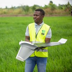 black man with drone in a green field - he's wearing a high visibility jacket 