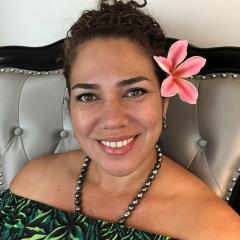 Coral Pasisi with a hibiscus flower behind her left ear, hair back, wearing necklance with bare shoulder dress - smiling to camera. 