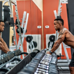 Black man with large, tattooed muscular arms leaning in to look at himself in a gym mirror. 