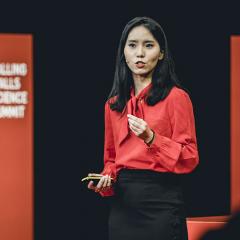 Dr Clara Jiang wearing a red shirt, and gesturing as she stands on stage speaking.