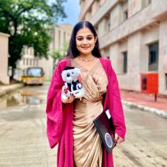 Dr Neha Singh standing in front of a building wearing a pink graduation gown. 