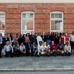 Group photo of all participants with banners of Australia Awards and UQ International Development on either side. The large group is standing in front of a brick wall with old fashioned windows. 