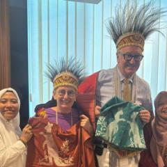 Ibu Tity with course leads Hon. Rachel Nolan and Professor Ken Smith AO wearing traditional Indonesian headdress - feathered - and holding Australian t-shirts with kangaroos 