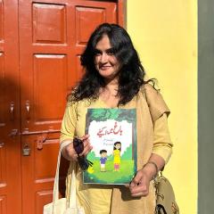 Saima Mazhar standing in front of a red door holding her published book. 