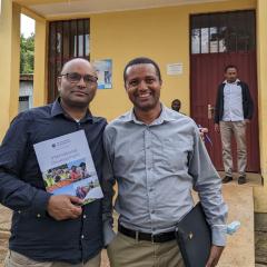 Yibeltal and PHCE staff Eskinder at health post in Ethiopia smiling to camera and holding up an International Development brochure