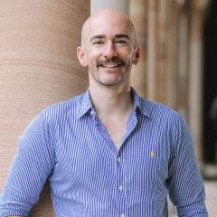 Adam smiling and standing in front of sandtone columns in UQ's Great Court