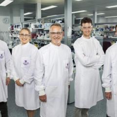 Five scientists smiling to camera wearing white lab coats with purple UQ insignia: man, woman, and three men including lead Professor Paul Young