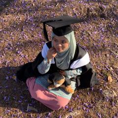 Nur Fazlini Ismail sitting on the ground in full graduation gown with a teddy bear also wearing a UQ graduation gown. Purple flowers from the Jacaranda tree are strewn around her. 