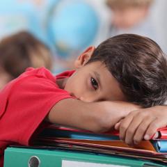Brown-skinned boy wearing red shirt laying his head on his arms over school desk looking to camera.