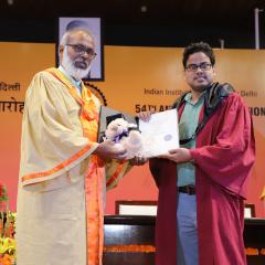 Dr Alok Kumar Ray holding PhD degree on stage with red robes and colourful orange flowers