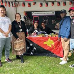 A group of people sitting and standing at a stall draped with a PNG  flag at UQ's Market Day, 2024