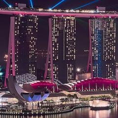 view of Singapore's marina bay, an area of skyscrapers, hotels and malls, at night with pink and blue lights.